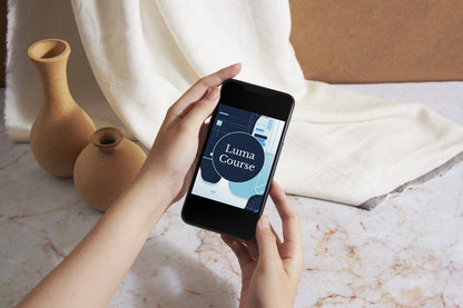 A person holds a smartphone showing a mockup or design labeled 'Luma Course' over a styled tabletop scene with cream fabric, two tan ceramic vases, and a brown backdrop.